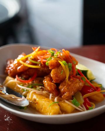 Crispy sweet-and-sour chicken with pineapple chunks, red and yellow bell peppers, bean sprouts and scallions, garnished with a lime wedge on a white restaurant plate