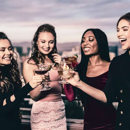 Four women toasting cocktails on a rooftop at sunset, laughing friends holding wine and mixed-drink glasses with a blurred city skyline backdrop.