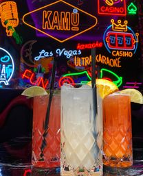 Three colorful cocktails in diamond-cut highball glasses with lime and orange garnishes and black straws, set against vibrant Las Vegas–style neon signage for karaoke, casino and fun.