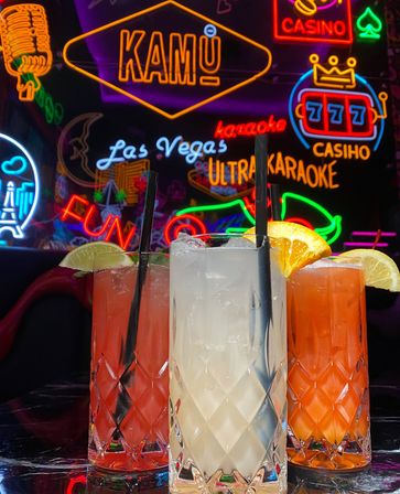 Three colorful cocktails in diamond-cut highball glasses with lime and orange garnishes and black straws, set against vibrant Las Vegas–style neon signage for karaoke, casino and fun.