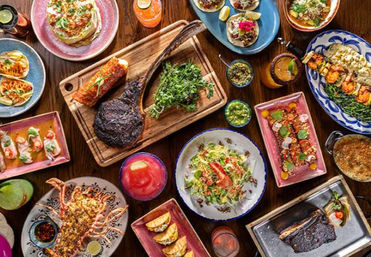 Vibrant table spread of grilled steak, shrimp skewers, tacos, small plates, fresh salads, sauces and colorful cocktails on a wooden table