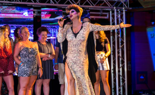 Showstopping drag performer in a money-print gown sings into a microphone on a brightly lit club stage, arm outstretched as smiling audience members stand behind during a lively drag show.