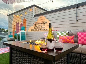 Cozy urban rooftop patio at dusk — lit fire-pit table with a wine bottle and two glasses of red, bright patterned cushions on a sectional, string lights overhead, and a painted city skyline mural with a handwritten quote on the wall.