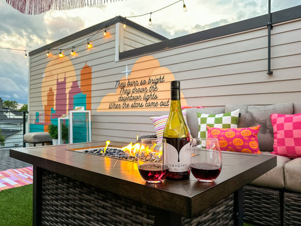 Cozy urban rooftop patio at dusk — lit fire-pit table with a wine bottle and two glasses of red, bright patterned cushions on a sectional, string lights overhead, and a painted city skyline mural with a handwritten quote on the wall.