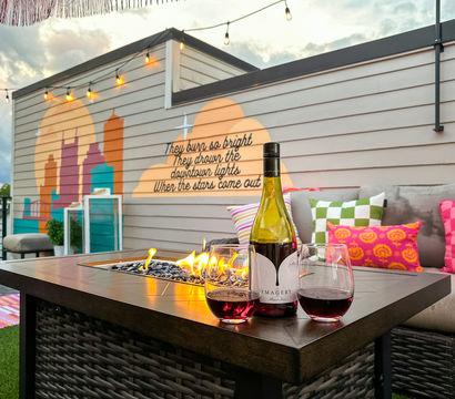 Cozy urban rooftop patio at dusk — lit fire-pit table with a wine bottle and two glasses of red, bright patterned cushions on a sectional, string lights overhead, and a painted city skyline mural with a handwritten quote on the wall.