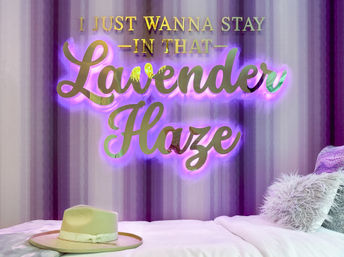 Lavender-themed bedroom with glowing gold script sign reading 'I just wanna stay in that Lavender Haze' backlit in purple, striped lavender wall, white bed with fluffy throw pillows and a beige wide-brim hat.