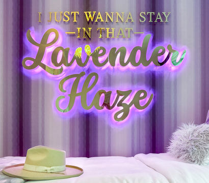 Lavender-themed bedroom with glowing gold script sign reading 'I just wanna stay in that Lavender Haze' backlit in purple, striped lavender wall, white bed with fluffy throw pillows and a beige wide-brim hat.