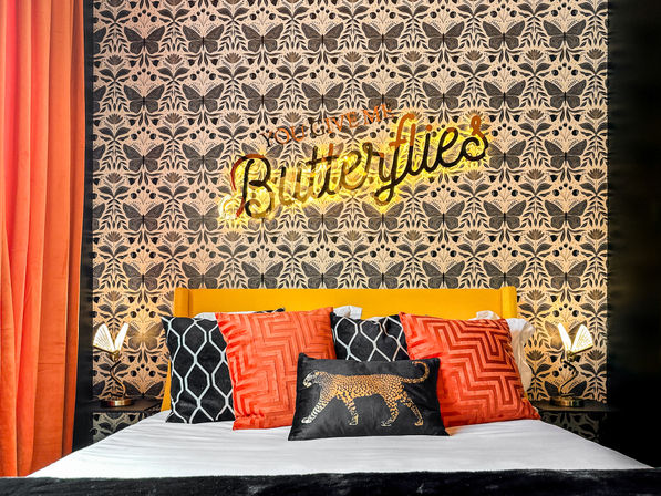 Stylish boutique bedroom with butterfly-patterned wallpaper and an illuminated Butterflies neon sign above a yellow headboard, bed dressed with orange and black geometric pillows and a leopard-print accent cushion, flanked by brass bedside lamps and orange velvet curtains.