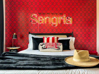 Playful boutique bedroom with red patterned wallpaper and illuminated “Sangria” sign above a neatly made bed, black throw, striped and embroidered pillows, bedside lamp and a beige wide‑brim hat on the foot of the bed.