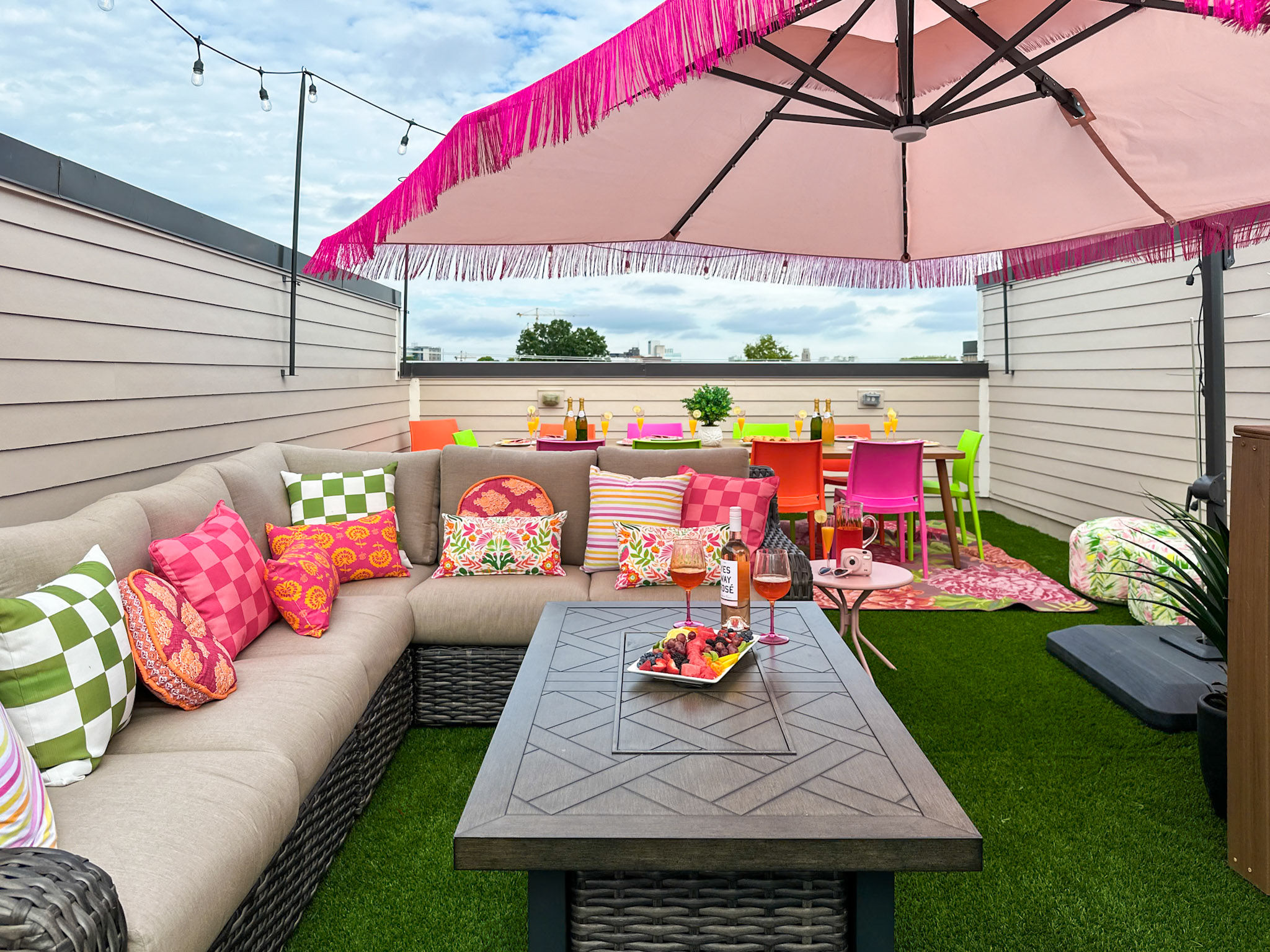 Vibrant urban rooftop patio with beige sectional sofa and colorful patterned pillows, pink fringed umbrella, wicker coffee table with fruit platter and rosé, bright neon dining chairs on artificial turf under string lights