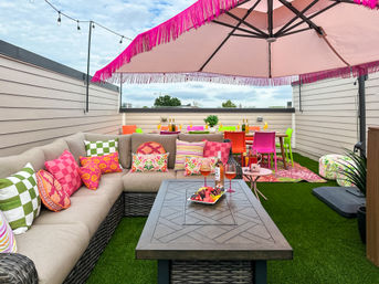 Vibrant urban rooftop patio with beige sectional sofa and colorful patterned pillows, pink fringed umbrella, wicker coffee table with fruit platter and rosé, bright neon dining chairs on artificial turf under string lights