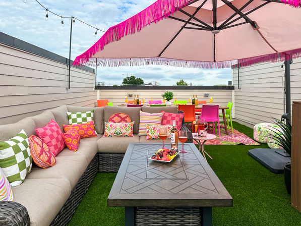 Vibrant urban rooftop patio with beige sectional sofa and colorful patterned pillows, pink fringed umbrella, wicker coffee table with fruit platter and rosé, bright neon dining chairs on artificial turf under string lights