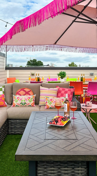 Vibrant urban rooftop patio with beige sectional sofa and colorful patterned pillows, pink fringed umbrella, wicker coffee table with fruit platter and rosé, bright neon dining chairs on artificial turf under string lights