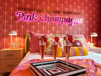 Three rosé champagne flutes with strawberry garnishes on a striped tray on a bed in a pink retro bedroom with patterned wallpaper and a neon “Pink Champagne” sign