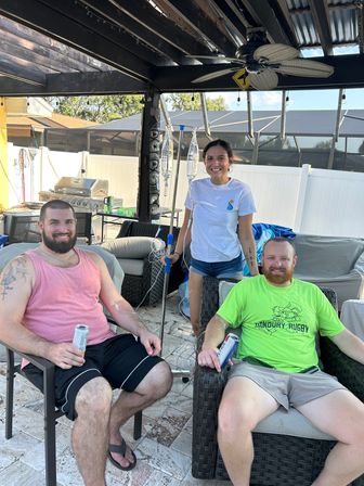 Three adults smiling on a sunny backyard patio with a pergola and screened pool enclosure — two men seated with drinks while a woman stands holding an IV pole with hanging saline bags.