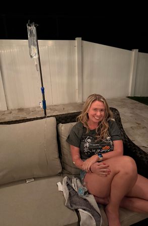 Smiling woman seated on a wicker outdoor sofa in a backyard patio at night, wearing a skeleton Halloween t‑shirt and smartwatch, with an IV drip bag hanging from a pole beside her, beige cushions and a white privacy fence in the background.