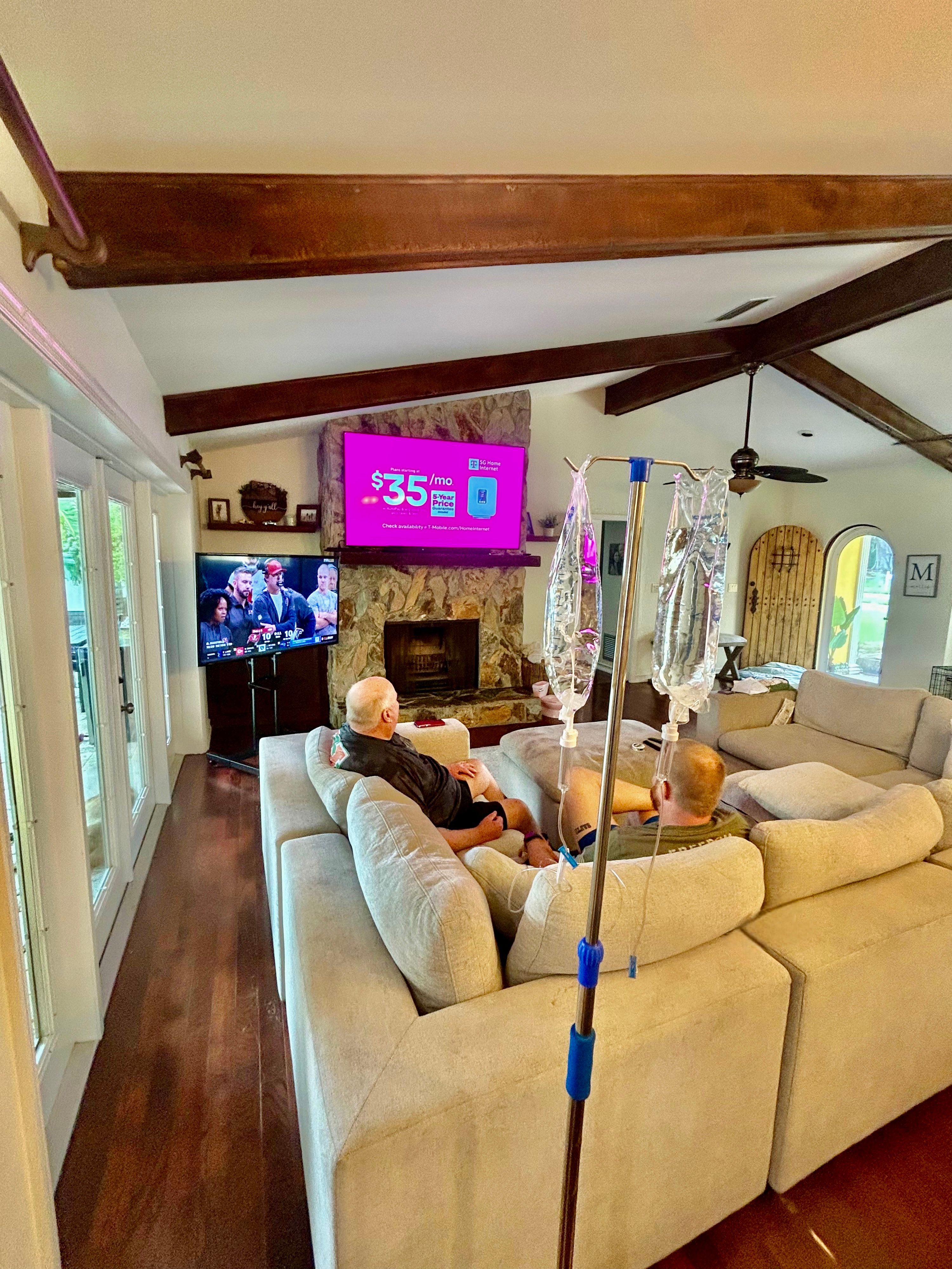 Cozy home living room with exposed wooden beams and hardwood floors, beige sectional sofa with two people watching two TVs over a stone fireplace, IV pole with hanging IV bags in the foreground, glass doors and arched wooden entry visible