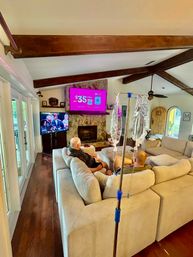 Cozy home living room with exposed wooden beams and hardwood floors, beige sectional sofa with two people watching two TVs over a stone fireplace, IV pole with hanging IV bags in the foreground, glass doors and arched wooden entry visible