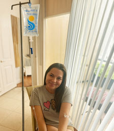 Smiling woman sitting in a sunlit living room by sliding glass doors with vertical blinds, receiving an IV infusion from a hanging saline bag on a pole with tubing taped to her arm, wearing a gray t-shirt with a heart and leopard-print patch.