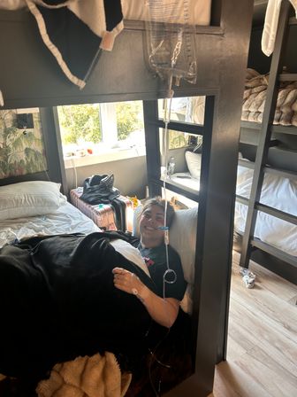 Smiling person lying in a lower bunk bed in a sunlit hostel-style room, covered with a black blanket and hooked up to an IV drip from a hanging bag; suitcase, pillows and cozy bedding visible.