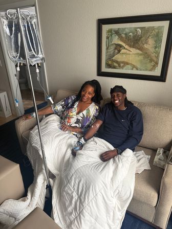 Smiling couple on a beige sofa receiving IV infusion therapy from hanging bags on a pole, wrapped in white blankets beneath a framed bird painting in a cozy room.