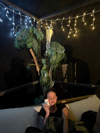 Woman relaxing on a cozy screened porch couch at night under twinkling string lights, eyes closed holding a small card, with a large leafy plant and an IV drip bag hanging from a pole nearby.