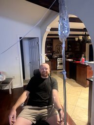 Smiling man in shorts sitting in a home kitchen receiving an at-home IV infusion from a hanging bag and tubing, with arched doorway, dark wood beams and tiled floor visible.