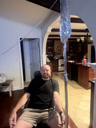 Smiling man in shorts sitting in a home kitchen receiving an at-home IV infusion from a hanging bag and tubing, with arched doorway, dark wood beams and tiled floor visible.