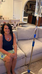 Smiling woman seated on a beige couch in a cozy home living room receiving an IV infusion from a pole, dining table and kitchen visible in background.