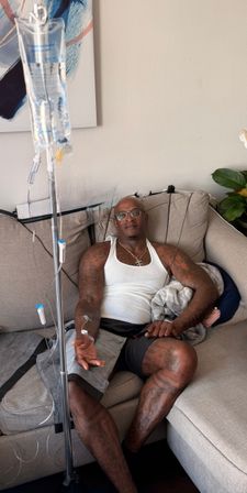 Tattooed man in a white tank top and glasses reclining on a beige living-room sofa while receiving an IV drip; clear IV bag and tubing, abstract wall art and potted plant in the background.