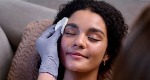 Relaxed woman with eyes closed receiving a spa facial cleansing from a gloved esthetician using a cotton pad — skincare treatment.