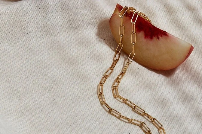 Gold paper-clip chain necklace draped over a ripe peach slice on textured cream fabric