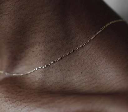 Close-up of a dark-skinned neck and collarbone with a delicate gold chain, minimalist jewelry portrait showcasing textured skin and soft studio lighting.