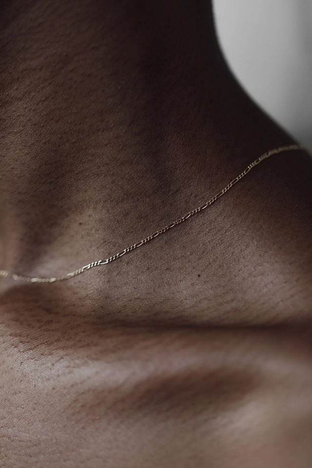 Close-up of a dark-skinned neck and collarbone with a delicate gold chain, minimalist jewelry portrait showcasing textured skin and soft studio lighting.