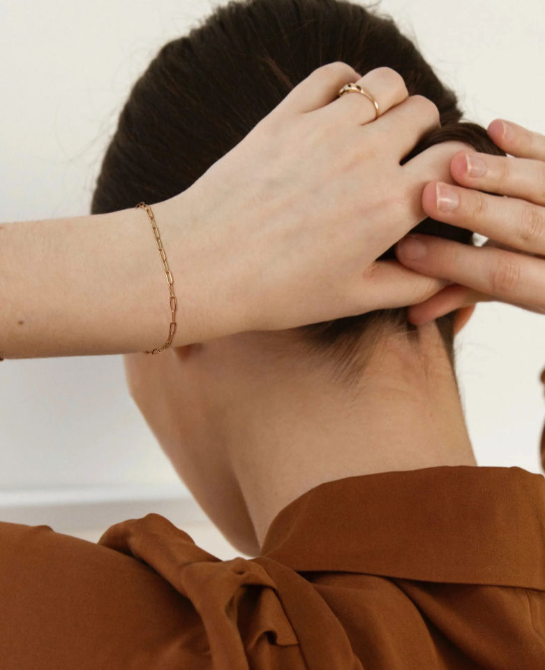 Close-up rear view of a person tying hair into a neat bun indoors, showcasing a delicate gold chain bracelet, slim gold ring, and a rust-brown blouse with a tied collar — minimalist fashion detail shot.