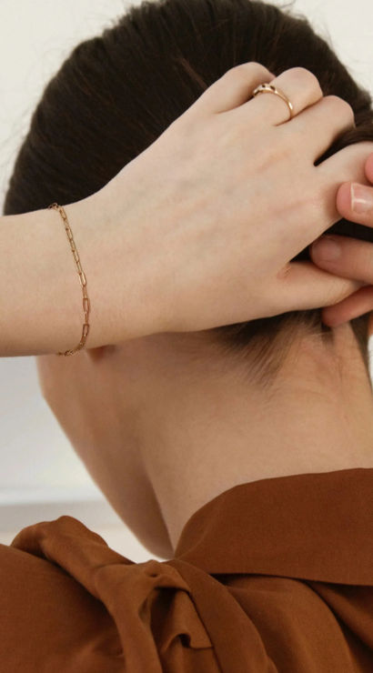 Close-up rear view of a person tying hair into a neat bun indoors, showcasing a delicate gold chain bracelet, slim gold ring, and a rust-brown blouse with a tied collar — minimalist fashion detail shot.