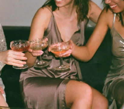 Three women in satin and sequined dresses toasting with cocktail coupe glasses while seated on a lounge couch at an indoor party