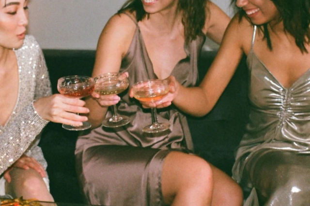 Three women in satin and sequined dresses toasting with cocktail coupe glasses while seated on a lounge couch at an indoor party