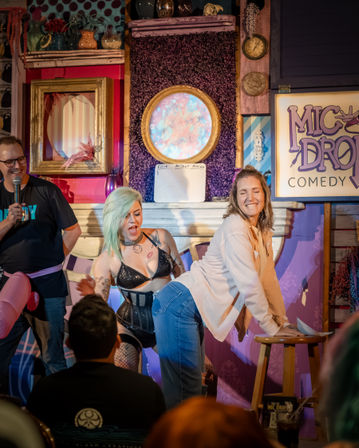 Energetic comedy club stage interior with a host holding a microphone, a burlesque-style performer in black and a laughing participant leaning on a stool amid colorful, retro decor and an attentive audience.