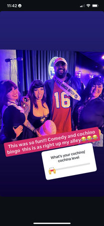 Group photo in a comedy club at a lively bingo night — four people under purple neon lights, a man wearing a "Bad Bingo Bitch" sash and #16 jersey holding an inflatable football, two women in playful outfits smiling on stage with mic and curtain backdrop.
