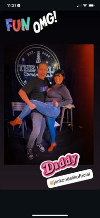 Playful photo on a comedy-club stage: a man lifts a smiling woman in jeans and heeled boots under colorful stage lights with 'FUN' and 'OMG' stickers.
