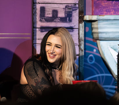 Smiling person with half-black, half-blonde hair and facial piercings in a fishnet top, laughing onstage against a colorful graffiti and wood-paneled backdrop with a microphone and red cup nearby