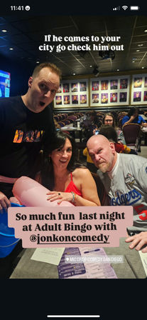 Three adults posing and laughing at a crowded comedy-club bingo night in San Diego — a woman in a red top holds a pink inflatable while two men make playful surprised faces, rows of tables and framed headshot photos fill the background.