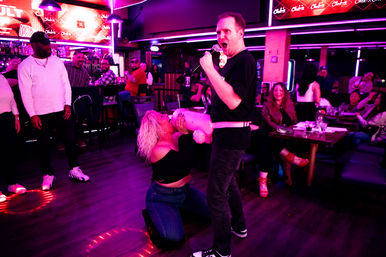 Neon-lit karaoke bar scene: singer with microphone and inflatable doll entertains a laughing crowd at tables.