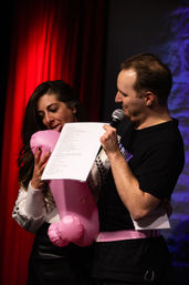 Two performers on stage in front of a red curtain — a man holds a microphone and script while a woman playfully bites a pink inflatable prop during a live comedy-style performance.