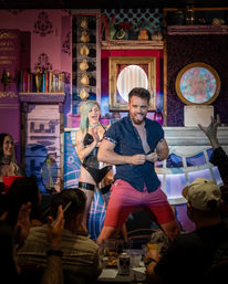 Man playfully dancing on a colorful cabaret stage with a costumed female performer and a cheering nightclub audience.