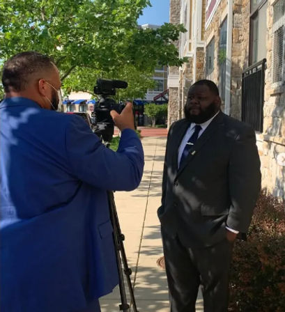 Outdoor downtown interview on a sunny sidewalk — a cameraman in a blue suit and face mask films a man in a dark suit and tie with a tripod-mounted video camera in front of a stone building and leafy trees.