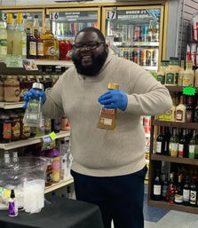 Smiling man in a sweater and blue gloves holding two liquor bottles at a liquor store tasting table, with shelves and refrigerated coolers of spirits and wine in the background.