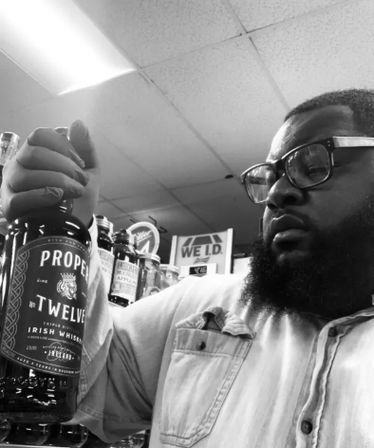 Person with glasses and beard examining a bottle of Irish whiskey in a liquor store aisle, retail shelves and a 'We I.D.' sign visible in the background