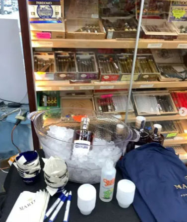 Retail cigar shop display with glass humidor of boxed cigars on shelves and a front table showing an ice bucket with a chilled bottle, disposable cups, napkins, pens and a navy promotional tote.